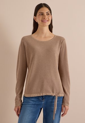 Kvinde med langt mørkt hår iført en lysebrun langærmet sweater med snørrelukning i kanten og blå jeans, smilende og kigger til siden.