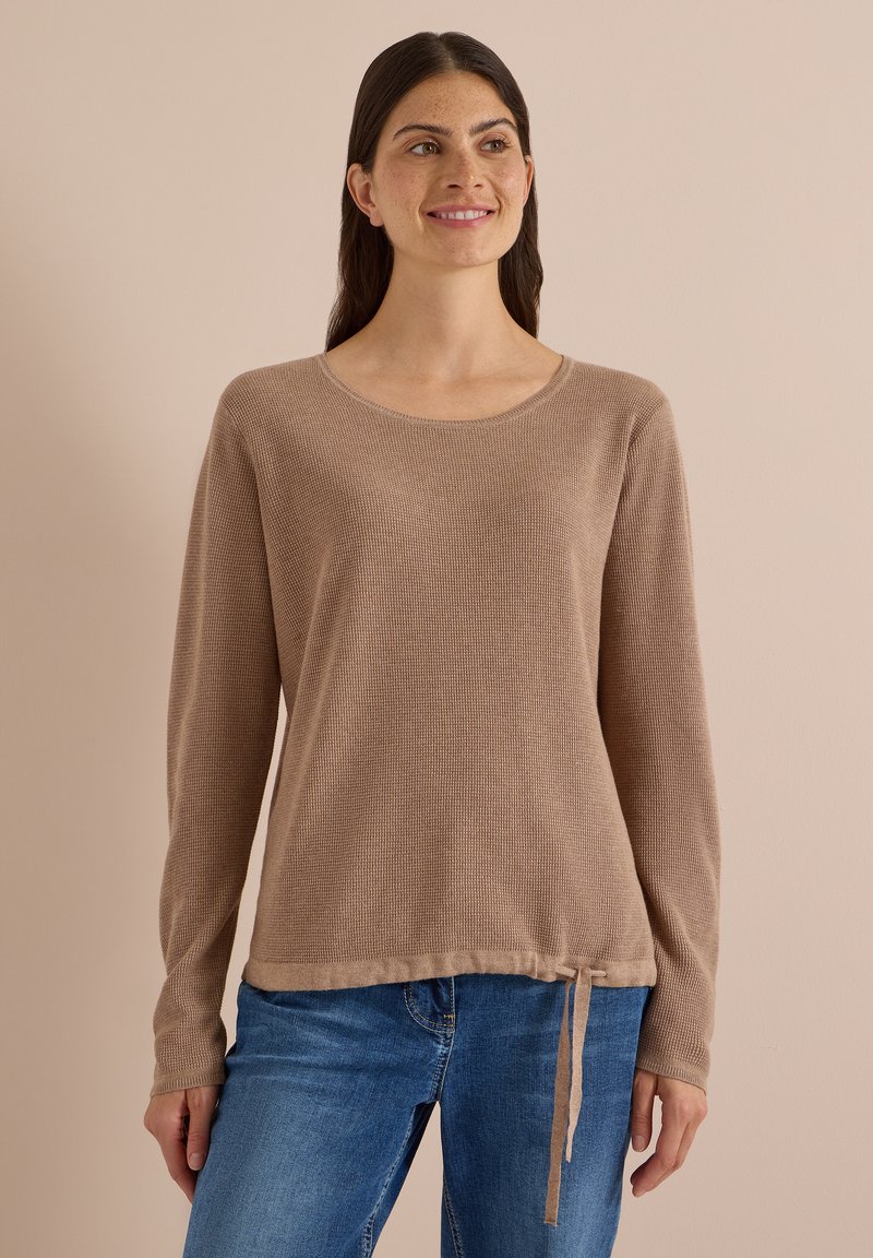 Femme aux longs cheveux foncés portant un pull marron clair à manches longues avec un ourlet à cordon de serrage et un jean bleu, souriante et regardant sur le côté.