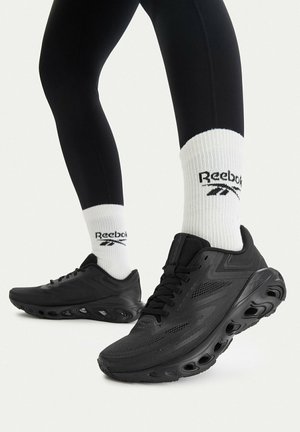 Person mit schwarzen Sportschuhen mit geformten Sohlen und weißen Reebok-Socken, im Halbgang vor einem schlichten hellen Hintergrund.