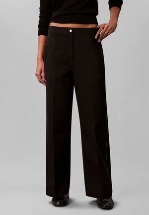 Persona che indossa pantaloni neri a vita alta e gamba larga con bottoni e tasche sul davanti, abbinati a scarpe nere e un top nero corto.