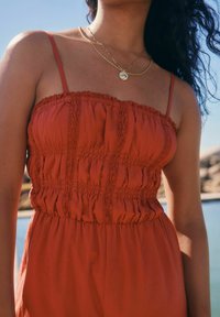 Robe couleur rouille avec un corsage smocké, fines bretelles et détails en dentelle. Texture lisse avec une coupe décontractée, sur fond d'eau.