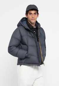 Giubbotto imbottito di colore blu scuro con cuciture a vista, chiusura con zip e cappuccio. Presenta dettagli arancioni sulla zip ed è abbinato a una camicia a quadri e pantaloni chiari.