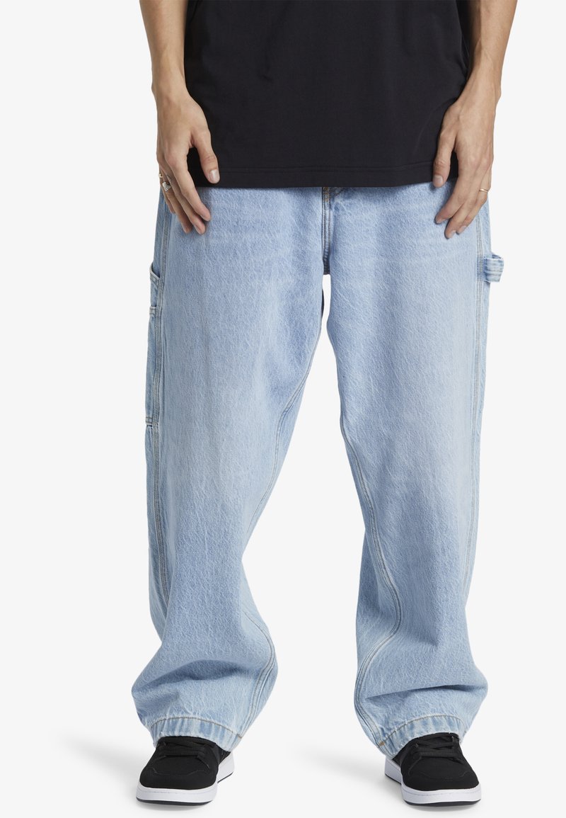 Helltürkise, übergroße Jeans aus Denim mit einer ausgewaschenen Textur, ausgestattet mit Seitentaschen und einem lockeren Schnitt, kombiniert mit schwarzen Sneakers.