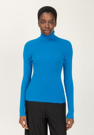 Een vrouw in een aansluitende felblauwe ribbelgebreide coltrui en ruime zwarte broek, staande tegen een effen lichte achtergrond.