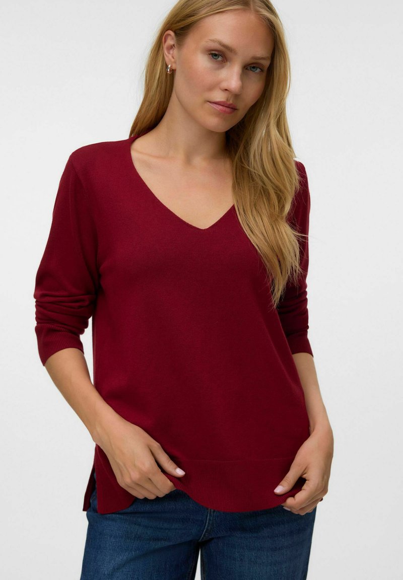 Femme aux longs cheveux blonds portant un pull rouge foncé à col en V et un jean bleu, debout devant un fond clair uni.