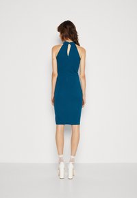 Türkisfarbenes, eng anliegendes, ärmelloses Kleid mit hohem Kragen und Schlüssellochrückendetail, aus glattem Stoff. Weiße High Heels und gemusterte Socken werden getragen.