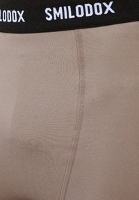 Close-up van beige stoffen ondergoed met zichtbare naad en een zwarte tailleband met witte SMILODOX-logo.