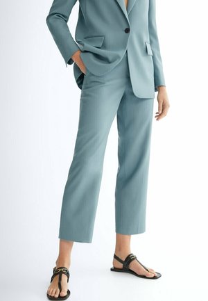 Vrouw in een lichtblauw op maat gemaakt pak met cropped broek en zwarte platte sandalen met gouden gesp, staand met één hand in de zak.