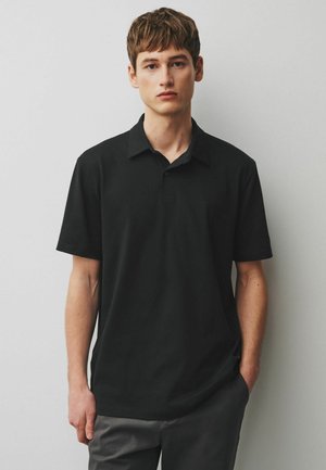 Nooremees lühikeste pruunide juustega, kes kannab musta lühikeste varrukatega polosärki, seisab ühetaustase helehalli seina taustal.