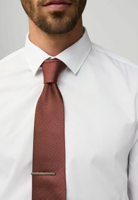 Červená textúrovaná kravata s úzkou striebornou sponou, nosená cez bielu košeľu s klasickým golierom. Detailný pohľad na oblasť krku.