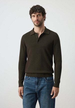 Hombre con cabello corto y rizado que lleva una camisa polo de manga larga color marrón oscuro y vaqueros azules, de pie frente a un fondo claro y liso.