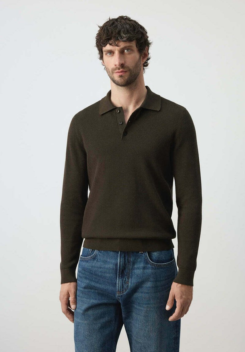 Homme aux cheveux courts et bouclés portant un polo marron foncé à manches longues et un jean bleu, debout devant un fond clair uni.