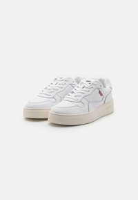 Białe sportowe sneakersy z teksturowaną cholewką, perforowanymi akcentami, płaskimi sznurówkami i kontrastującą beżową gumową podeszwą. Widoczny detal logo.