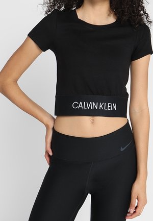 Kobieta ubrana w czarny krótki T-shirt Calvin Klein i czarne legginsy Nike, stojąca z jedną ręką na biodrze na jednolitym tle.