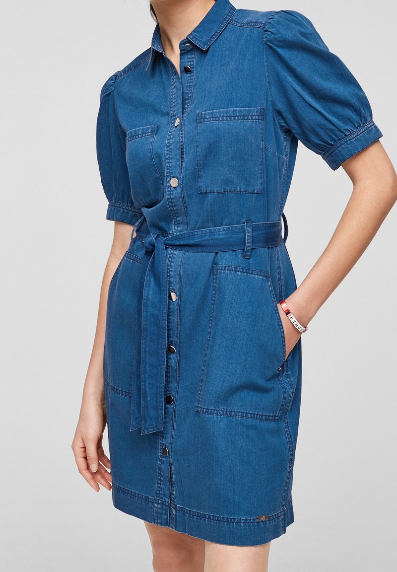 Robe chemise en denim bleu moyen, présentant des manches bouffantes, un devant à boutons, deux poches poitrine et une ceinture en tissu assortie.