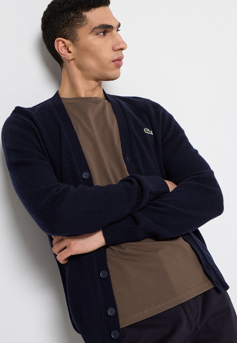 Giovane uomo con capelli corti e ricci che indossa un cardigan blu navy sopra una camicia marrone, in piedi con le braccia incrociate, che guarda di lato su uno sfondo bianco.