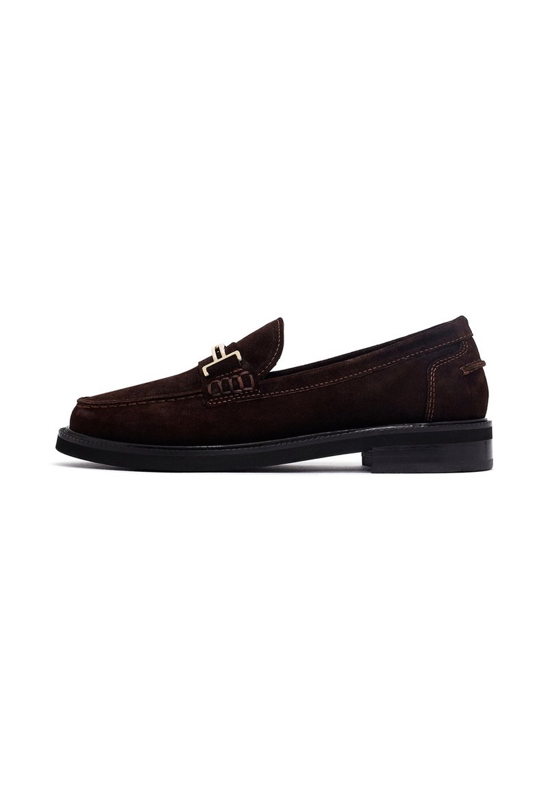 Mocasines de ante marrón con punta redondeada, tiras decorativas y acentos de hebilla, y suela de goma negra. Detalle de costura simple a lo largo de los lados.