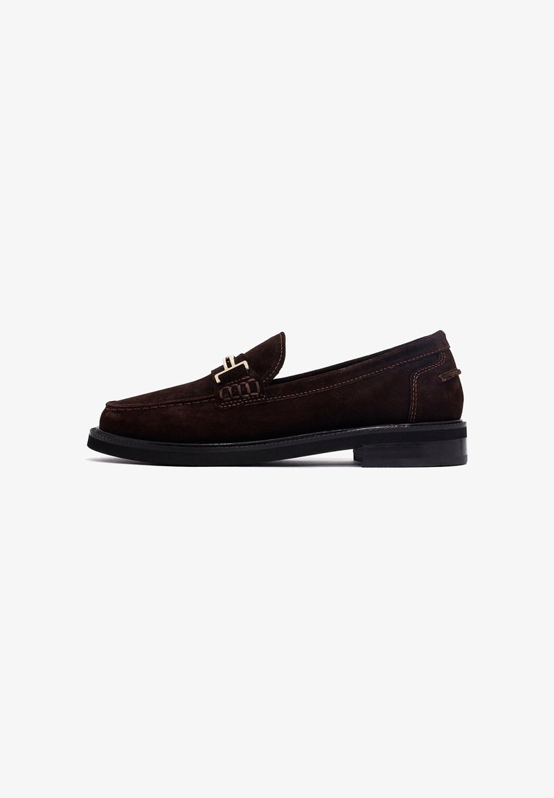 Mocassins en suede marron avec un bout rond, une sangle décorative et un accent de boucle, et une semelle en caoutchouc noire. Détails de couture simples le long des côtés.