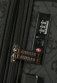 Bagaglio nero dalla texture particolare con doppie cerniere evidenziate da dettagli in oro con la scritta "GUESS?" e un pannello con serratura a combinazione con logo TSA.