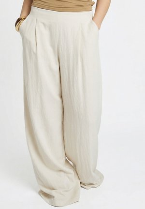 Vrouw draagt beige linnen broek met hoge taille en wijde pijpen, handen in de zakken, en een beige mouwloos topje, staand tegen een witte achtergrond.