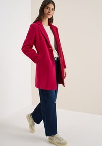 Fuchsiaroze wollen mix jas met een notchlapel, twee voorzakken, sluiting met één knoop, gecombineerd met donkerblauwe loszittende broeken en beige sneakers.