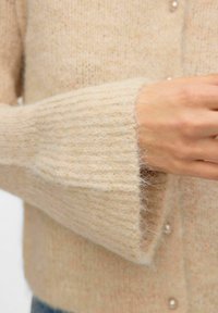 Gilet en laine beige avec des poignets côtelés, une texture douce et une fermeture à boutons en perles. Le tissu présente un motif subtil et moucheté.