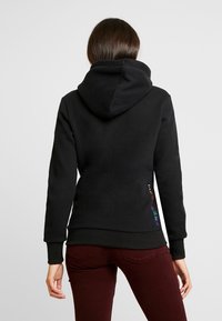 Černá mikina s kapucí s žebrovanými manžetami a lemem. Na boku jsou vícestranné vyšívané detaily. Měkký, strukturovaný materiál.