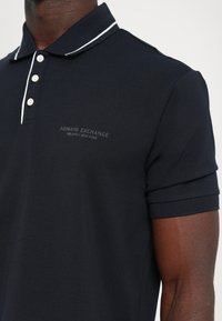 Marineblå polo t-skjorte med hvite detaljer på kragen. Inkluderer tre hvite knapper og merket "ARMANI EXCHANGE" på brystet. Glatt tekstur.
