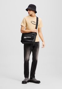 Bolso mensajero negro con logo blanco, llevado en bandolera. Modelo con camisa color beige claro, jeans grises y sombrero tipo bucket negro, de pie sobre un fondo gris.