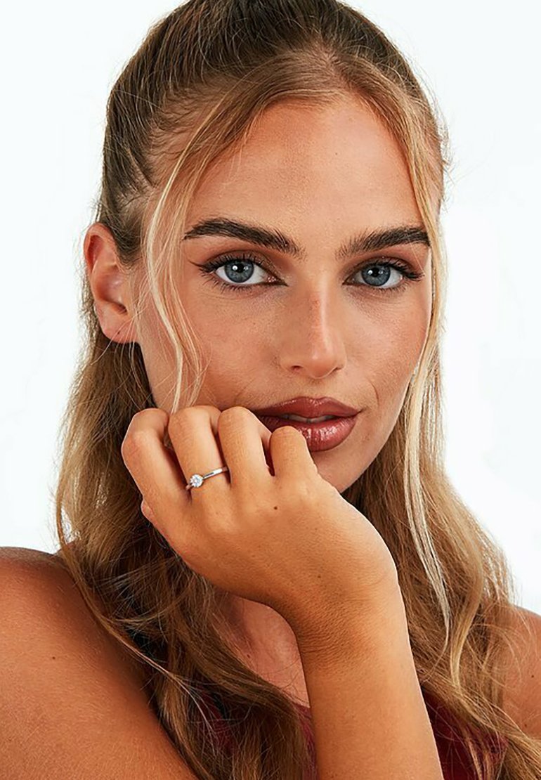 Silberring mit einem zentralen runden Diamanten, an der linken Hand getragen. Das Model hat lange, wellige blonde Haare und ist in einem Nahaufnahme-Porträt.