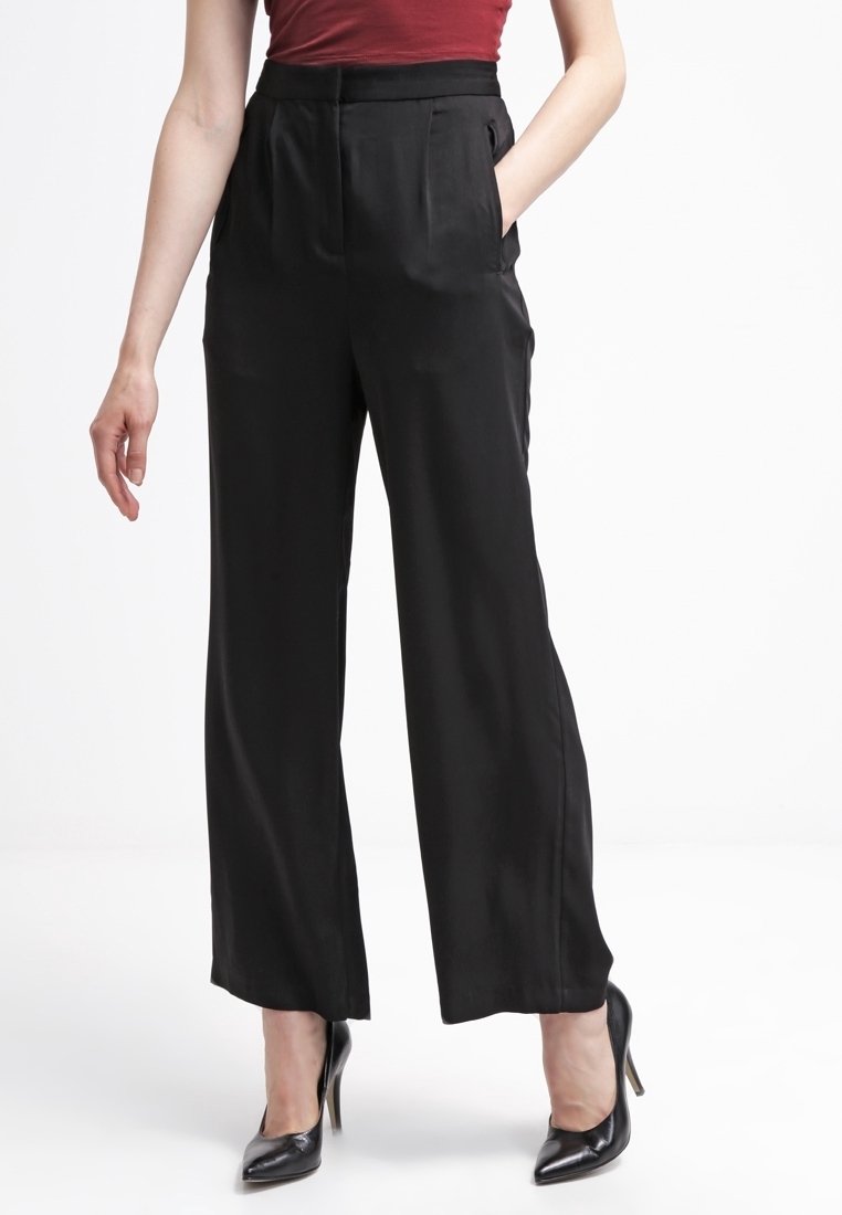Pantalon large en satin noir taille haute avec poches latérales, associé à des chaussures à talons noires, présentant une texture lisse.