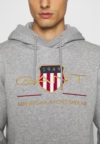 Grauer Kapuzenpullover mit strukturiertem Stoff, goldenen Akzenten und einem zentralen gestickten Emblem mit roten und blauen Streifen.
