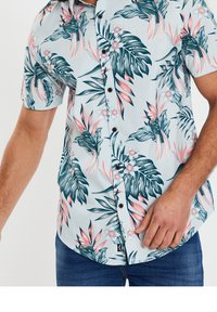 Chemise à manches courtes, à boutons, en tissu bleu clair, ornée d'un motif floral tropical avec des feuilles vertes et des accents roses. Boutons foncés visibles.