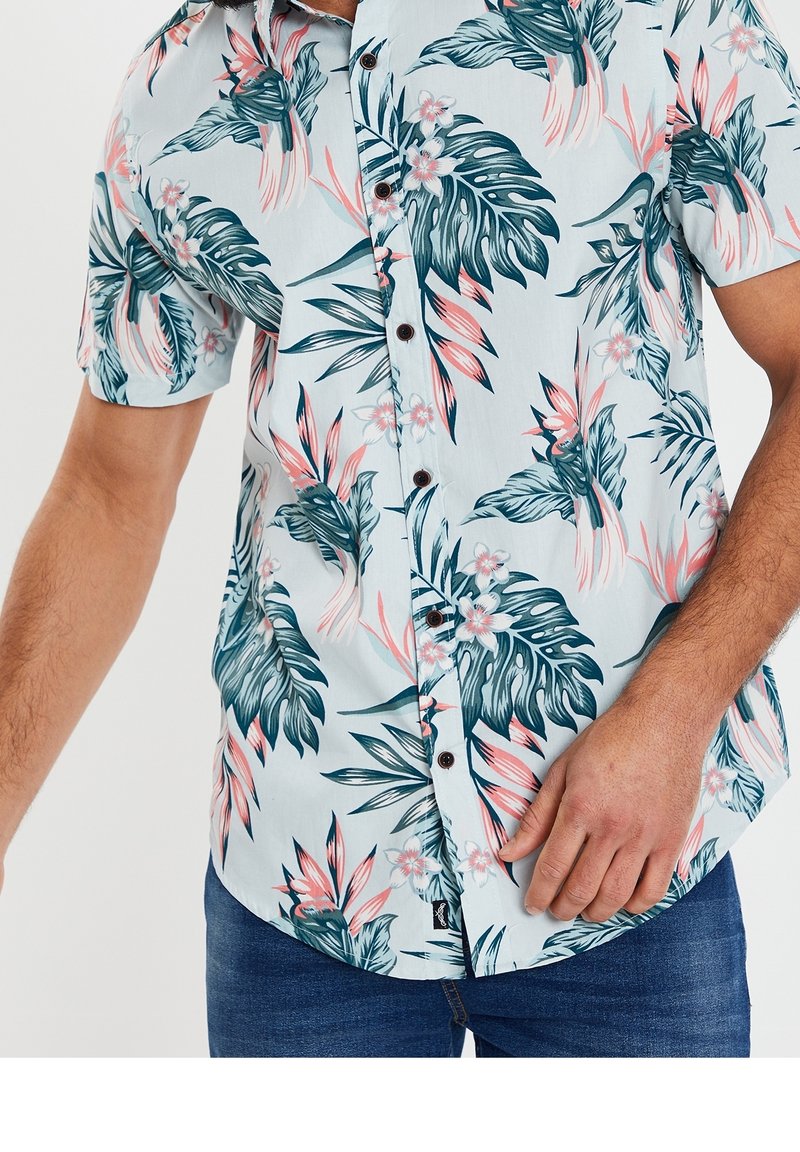 Chemise à manches courtes, à boutons, en tissu bleu clair, ornée d'un motif floral tropical avec des feuilles vertes et des accents roses. Boutons foncés visibles.