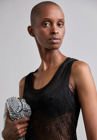 Top sin mangas de malla negra con un escote acanalado, con un diseño transparente. La modelo sostiene un accesorio plateado brillante con patrones intrincados.