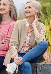 Zwei lächelnde ältere Frauen sitzen im Freien; die eine trägt einen beigen Cardigan und Jeans mit weißen Turnschuhen, die andere ein rosa gestreiftes Oberteil.