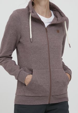 Osoba ubrana w fioletowawy mauve kaptur zapinany na zamek błyskawiczny z białymi sznurkami i kieszeniami kangurzymi, na białą koszulkę, ręce w kieszeniach.