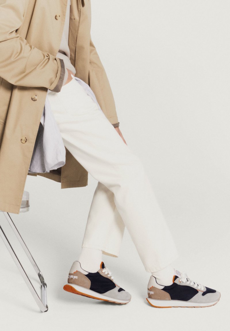Footwear featuring a mix of grey, navy, and tan suede, with orange accents, white laces, and a textured sole. Casual design with sporty elements.