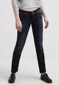 Jeans ajustados de mezclilla de color azul oscuro, con bolsillos delanteros, un ligero desgaste y costuras en contraste. Llevados con zapatos negros de cordones.