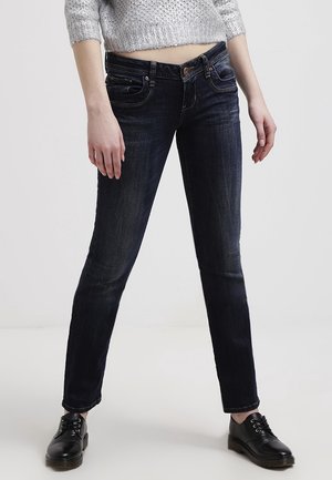 Jeans ajustados de mezclilla de color azul oscuro, con bolsillos delanteros, un ligero desgaste y costuras en contraste. Llevados con zapatos negros de cordones.