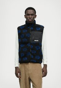 Fleece mellény kék leopárdmintával fekete alapon, magas gallérral, cipzáras mellkzsebbel, világos hosszú ujjú inggel párosítva.