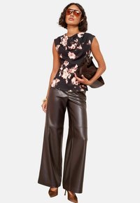 Top nero floreale con maniche a cappuccio, abbinato a pantaloni in pelle larghi di colore marrone scuro. Gli accessori includono una borsa marrone e scarpe con tacco marroni.