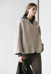 Frau trägt eine beige Reißverschlussjacke mit großen Taschen und dunkle Hosen, hält einen gefalteten schwarzen Mantel vor einem einfachen hellen Hintergrund.