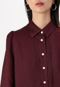 Burgundy blouse with a textured finish, featuring a collar, vertical pleats, and decorative pearl-like buttons.