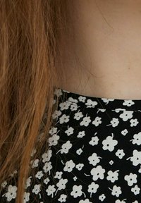 Svart tyg med små vita blommönster, med en rund halsringning och en strukturerad yta, kombinerat med lång vågig brunt hår.