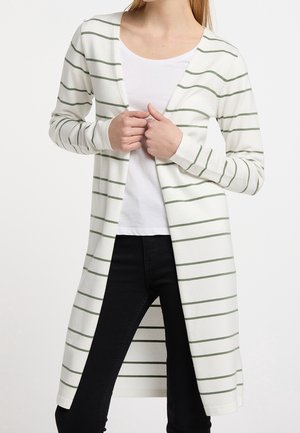 Femme portant un long cardigan blanc avec des rayures horizontales vertes, une chemise blanche et un pantalon noir, tenant le cardigan fermé au niveau de la poitrine.