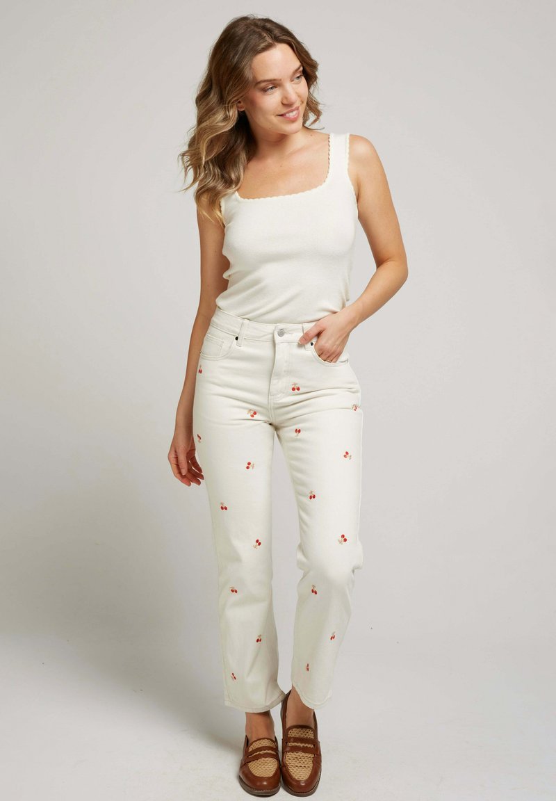 Débardeur blanc à côtes avec décolleté festonné, associé à un jean blanc orné de broderies en forme de cerise rouge. Des mocassins tissés bruns complètent le look.
