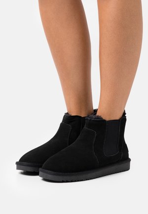 Bottes courtes en daim noir avec doublure en fourrure portées sur des jambes nues, dotées de panneaux latéraux élastiques et de semelles épaisses en caoutchouc.