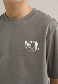 Pelēka kokvilnas T-kreklu ar apaļu apkakli un īsām piedurknēm. Iekļauj baltu tekstu "BLUE RIDGE" un grafiku ar sērfošanas dēļiem un viļņiem.
