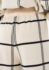 Beige gebreide broek met zwart geruit patroon. Heeft een zachte textuur en een elastische tailleband, met horizontale en verticale strepen.